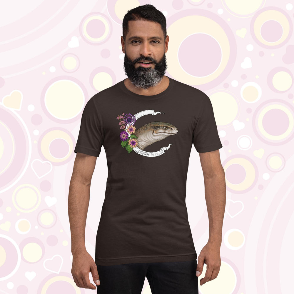 Man wearing brown crew neck tee with a head shot of a savannah monitor lizard with purple flowers on the side and a white banner encircling the design which says the species name.