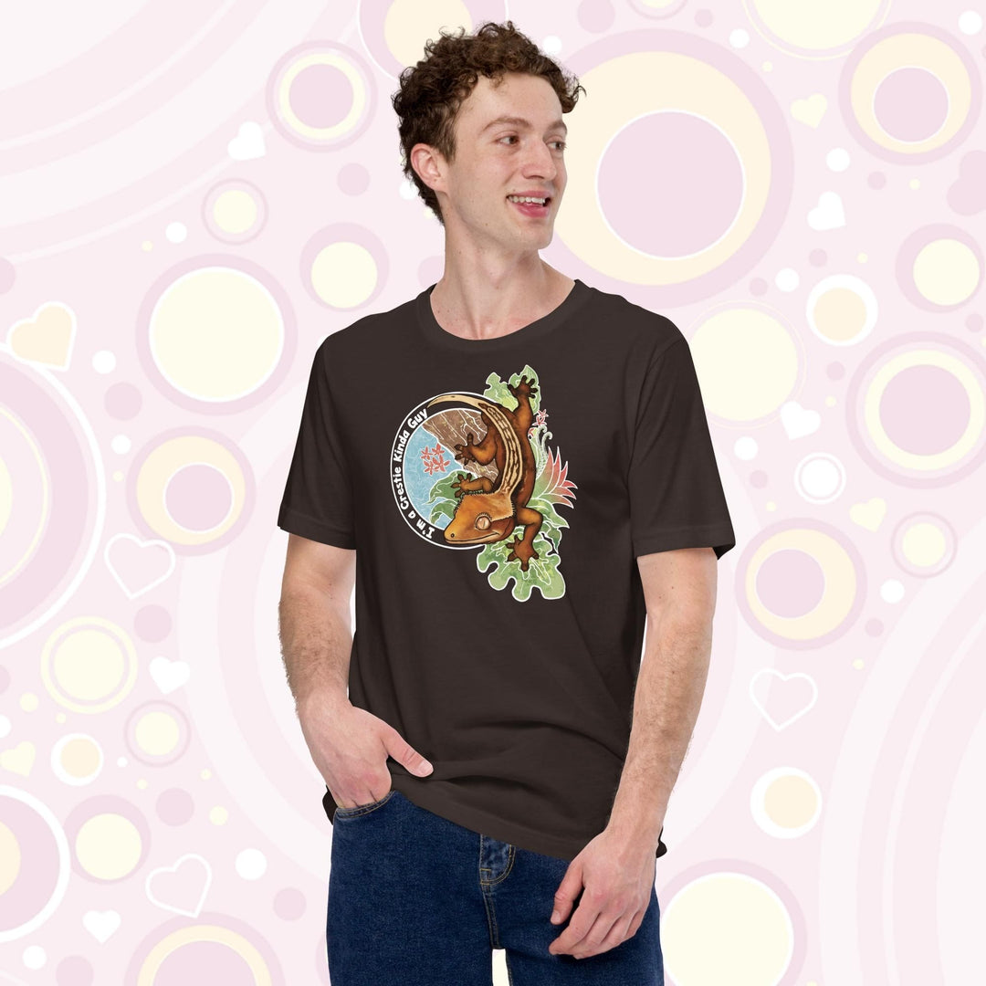 A man wearing a brown crew neck tee with a graphic of a crested gecko climbing down from a tree with the words "I'm a crestie kind of guy" on the left hand side.