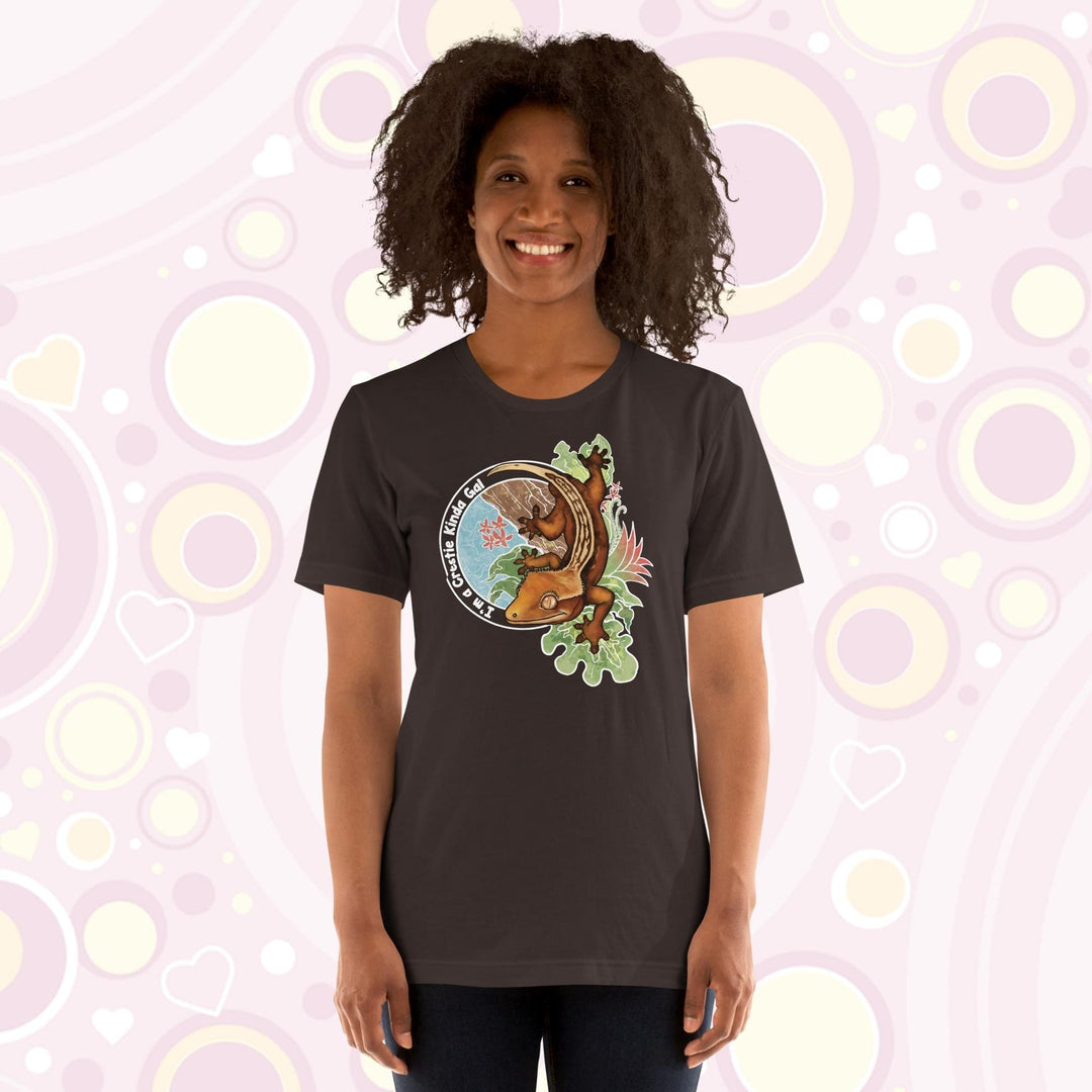 A woman wearing a brown crew neck tee with a graphic of a crested gecko climbing down from a tree with the words "I'm a crestie kind of gal" on the left hand side.