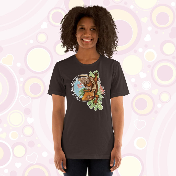 A woman wearing a brown crew neck tee with a graphic of a crested gecko climbing down from a tree with the words "I'm a crestie kind of gal" on the left hand side.