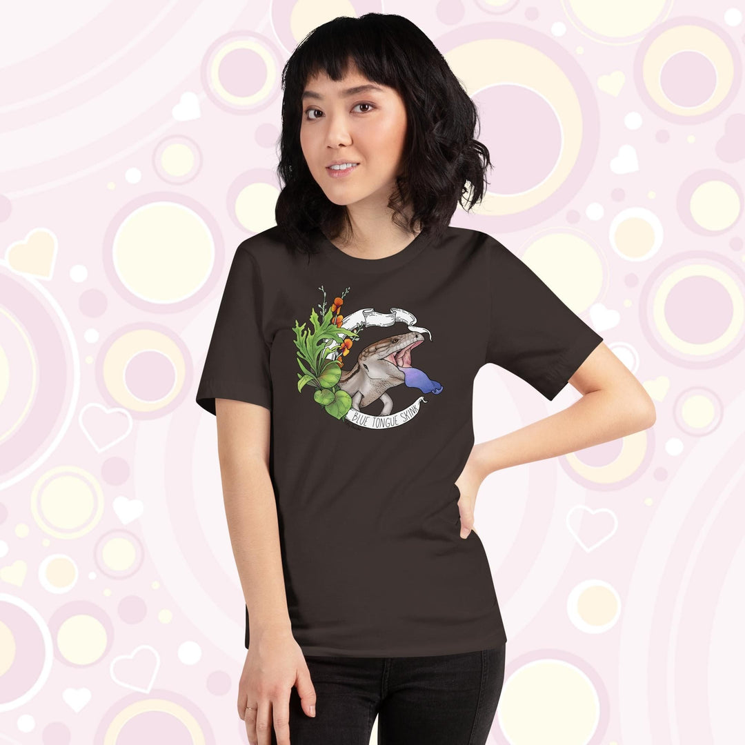 Lady wearing brown colored tee with a hand drawn design of a blue tongue skink flashing it's blue tongue in warning. Framed with greenery and a white banner that reads, "Blue Tongue Skink".