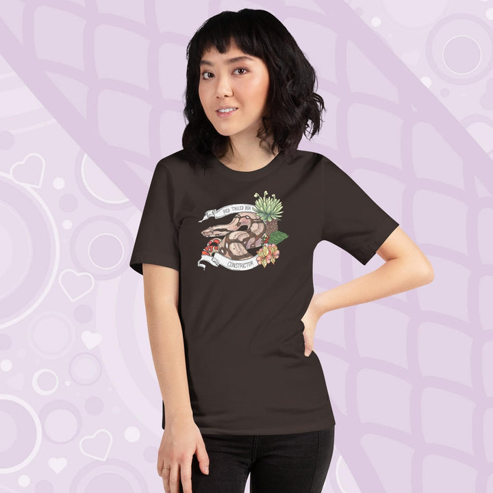 A woman wearing a brown crew necked tee with a graphic of a red-tailed boa constrictor with floral elements and a white banner encircling it. 
