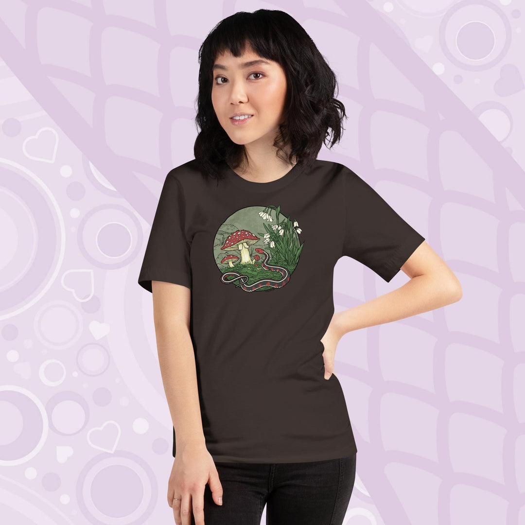 A woman wearing a brown tee shirt with a graphic of a red sided garter snake, surrounded by mushrooms and plants with white blossoms. It has simple colors of red, white, and green.  It has lineart outlining the entire illustration.