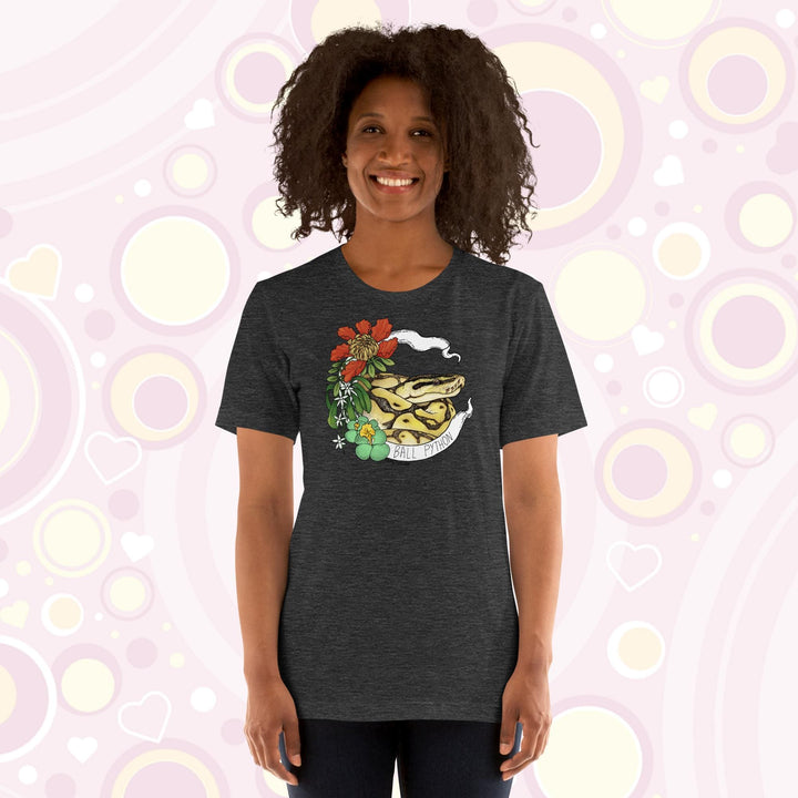 Image shows a woman wearing a heather grey crew neck tee with a ball python coiled up in a white banner with the snake species on it. Red, white, and green flowers and plants adorn the left side of the graphic.