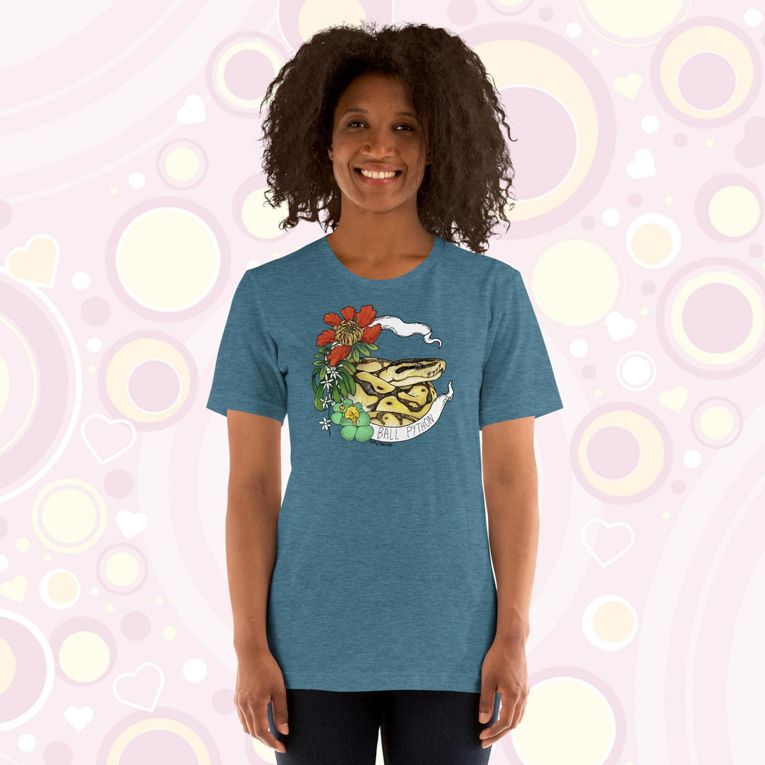 Image shows a woman wearing a heather teal crew neck tee with a ball python coiled up in a white banner with the snake species on it. Red, white, and green flowers and plants adorn the left side of the graphic.