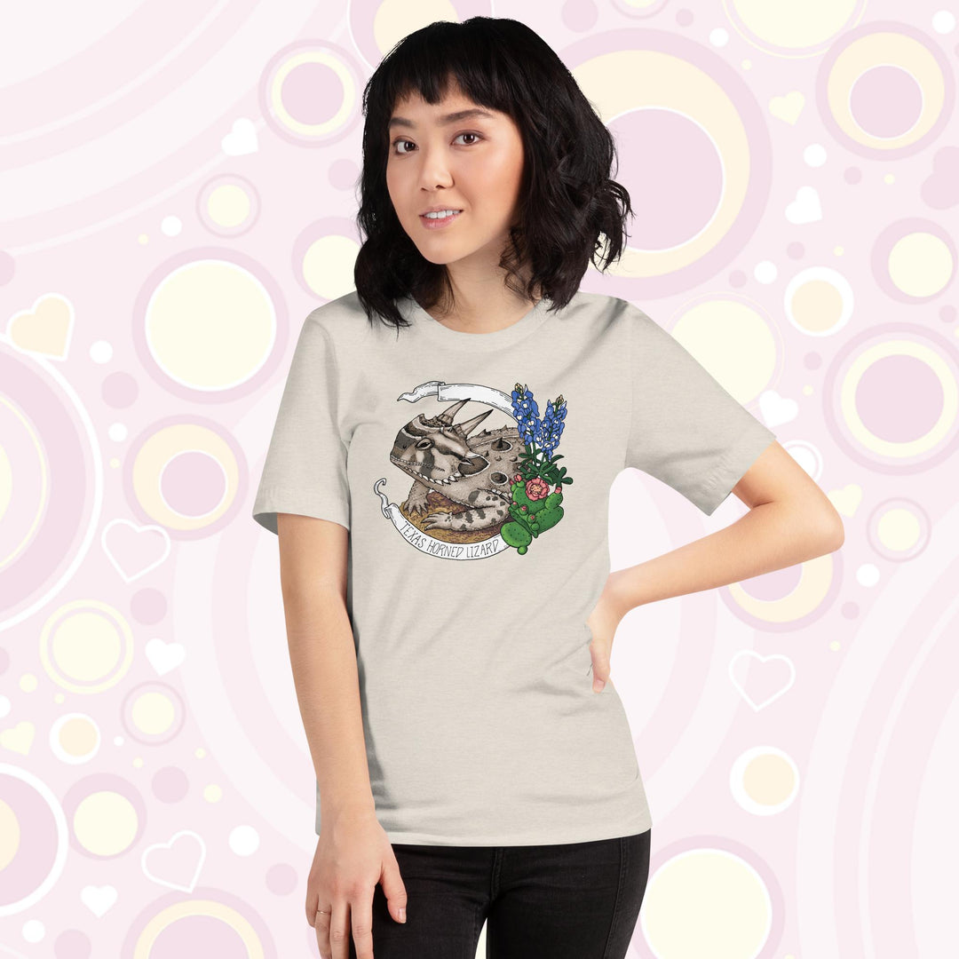 Woman wearing heather dust crew neck tee with a texas horn toad lizard resting on sand and framed by cactii and blue bonnet flowers. A banner encircles the design with the text "texas Horned Lizard" on it.