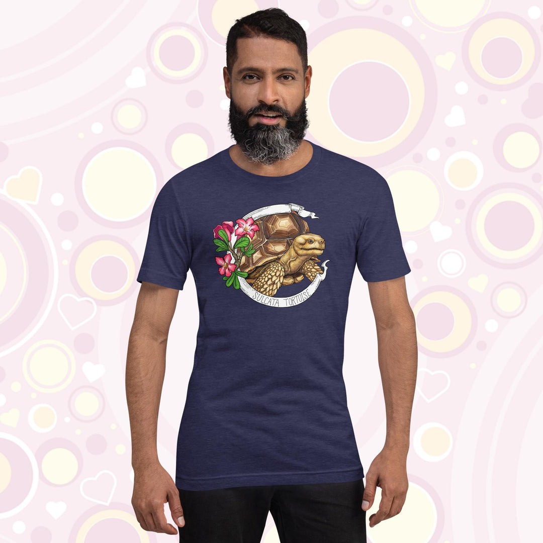 Man wearing heather midnight blue crew neck tee shirt with a sulcata tortoise in the middle of a white banner and pink flowers on the left side of the design. The banner reads Sulcata Tortoise.