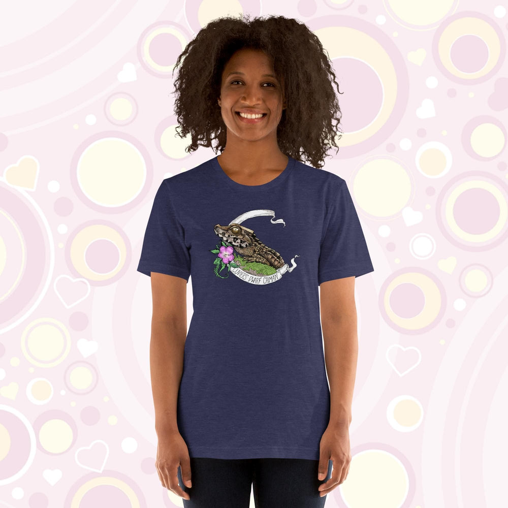 A woman wearing a heather midnight blue tee showing a Cuvier's Dwarf Caiman reptile perched on a mossy bed of greenery with a pink flower beneath it's head.