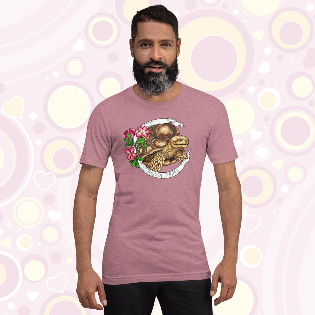 Man wearing mauve crew neck tee shirt with a sulcata tortoise in the middle of a white banner and pink flowers on the left side of the design. The banner reads Sulcata Tortoise.