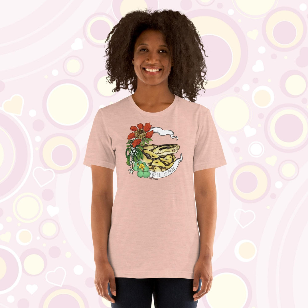 Image shows a woman wearing a heather peach crew neck tee with a ball python coiled up in a white banner with the snake species on it. Red, white, and green flowers and plants adorn the left side of the graphic.