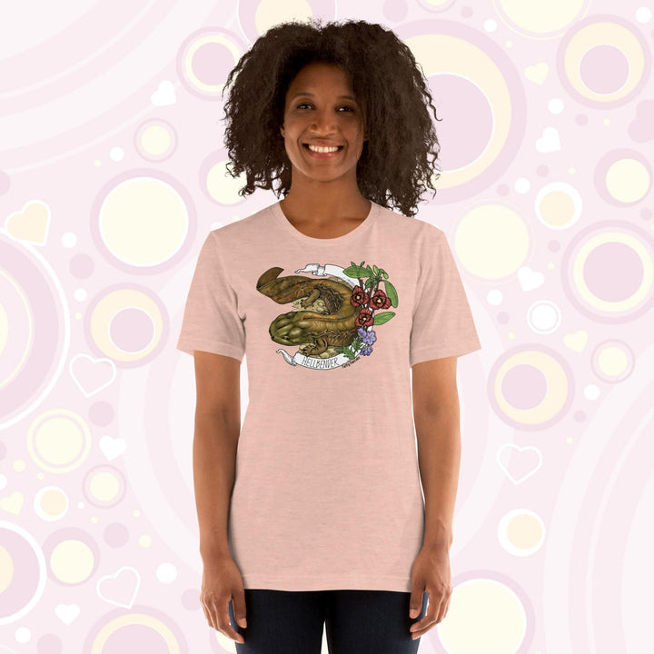 Lady wearing a peach crew neck tee with a hand drawn design of a hellbender amphibian framed by paw paw flowers and a banner that says, "Hellbender".