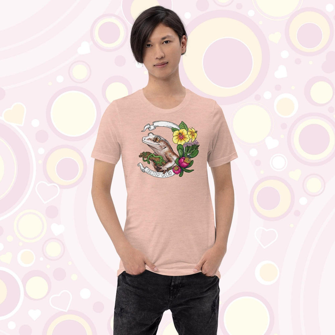 A teen wearing a peach crew neck tee of a crested gecko holding a branch with yellow hibiscus and other pink flowers as accents along the right of the design. A circular banner reads the name of the animal species on it.