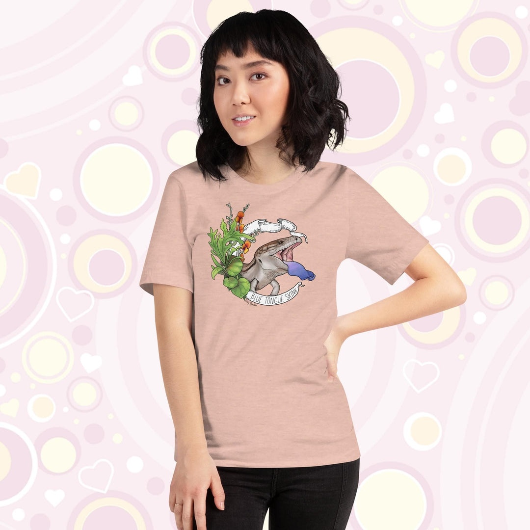 Lady wearing heather prism peach colored tee with a hand drawn design of a blue tongue skink flashing it's blue tongue in warning. Framed with greenery and a white banner that reads, "Blue Tongue Skink".