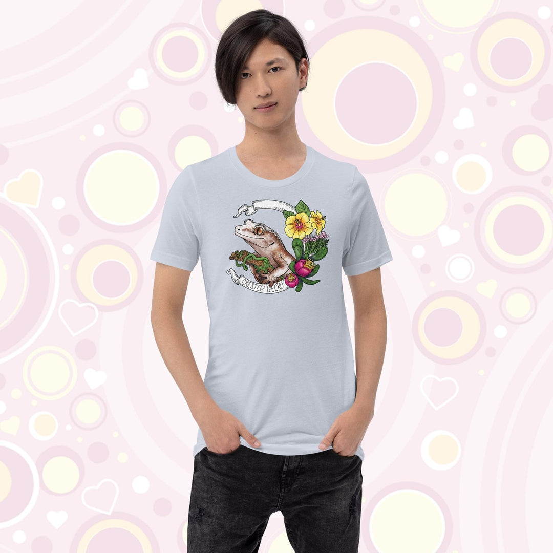 A teen wearing a light blue crew neck tee of a crested gecko holding a branch with yellow hibiscus and other pink flowers as accents along the right of the design. A circular banner reads the name of the animal species on it.