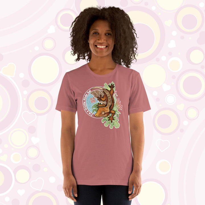 A woman wearing a mauve crew neck tee with a graphic of a crested gecko climbing down from a tree with the words "I'm a crestie kind of gal" on the left hand side.