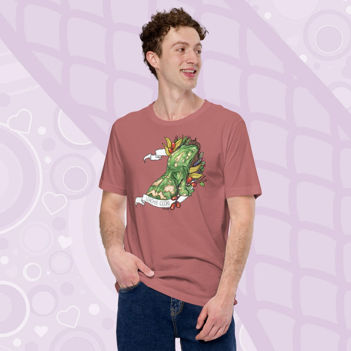 A guy wearing a neutrai pink colored tee with a green leachie gecko resting on a log and framed by green leaves and red berries. A white banner frames the reptile with the name "Leachie Gecko" on the bottom.