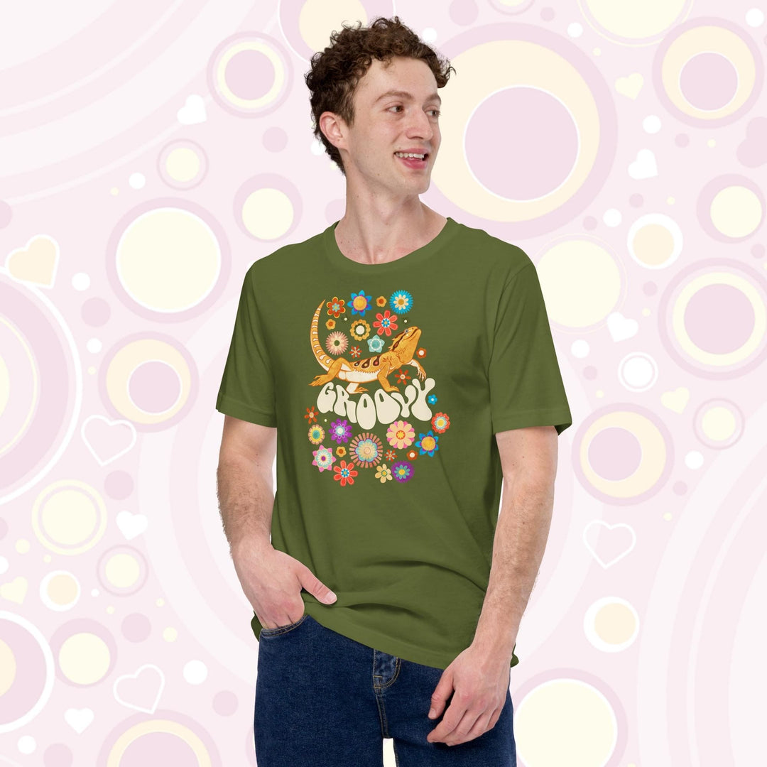 A young man wearing a olive crew neck tee showing a groovy bearded dragon design with a 70's retro style with many multi colored flowers surrounding a strutting bearded dragon lizard with the text "groovy" under it's body.