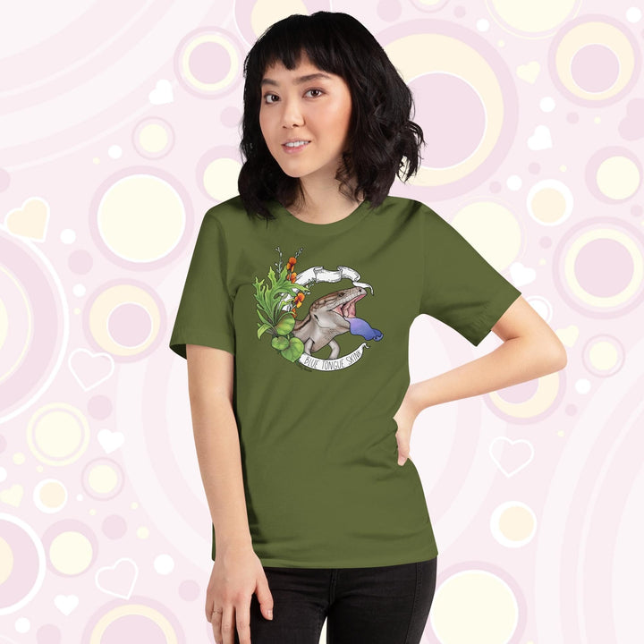 Lady wearing olive colored tee with a hand drawn design of a blue tongue skink flashing it's blue tongue in warning. Framed with greenery and a white banner that reads, "Blue Tongue Skink".