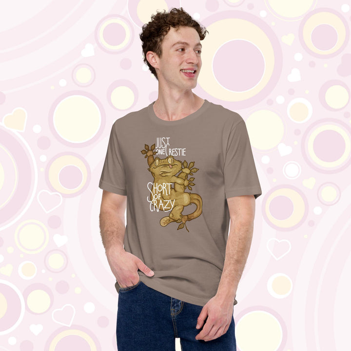 Man wearing pebble brown crew neck tee with the words "Just one Crestie Short of Crazy" with a crested gecko design clinging to the center. It's legs are flung out and it has a silly expression.