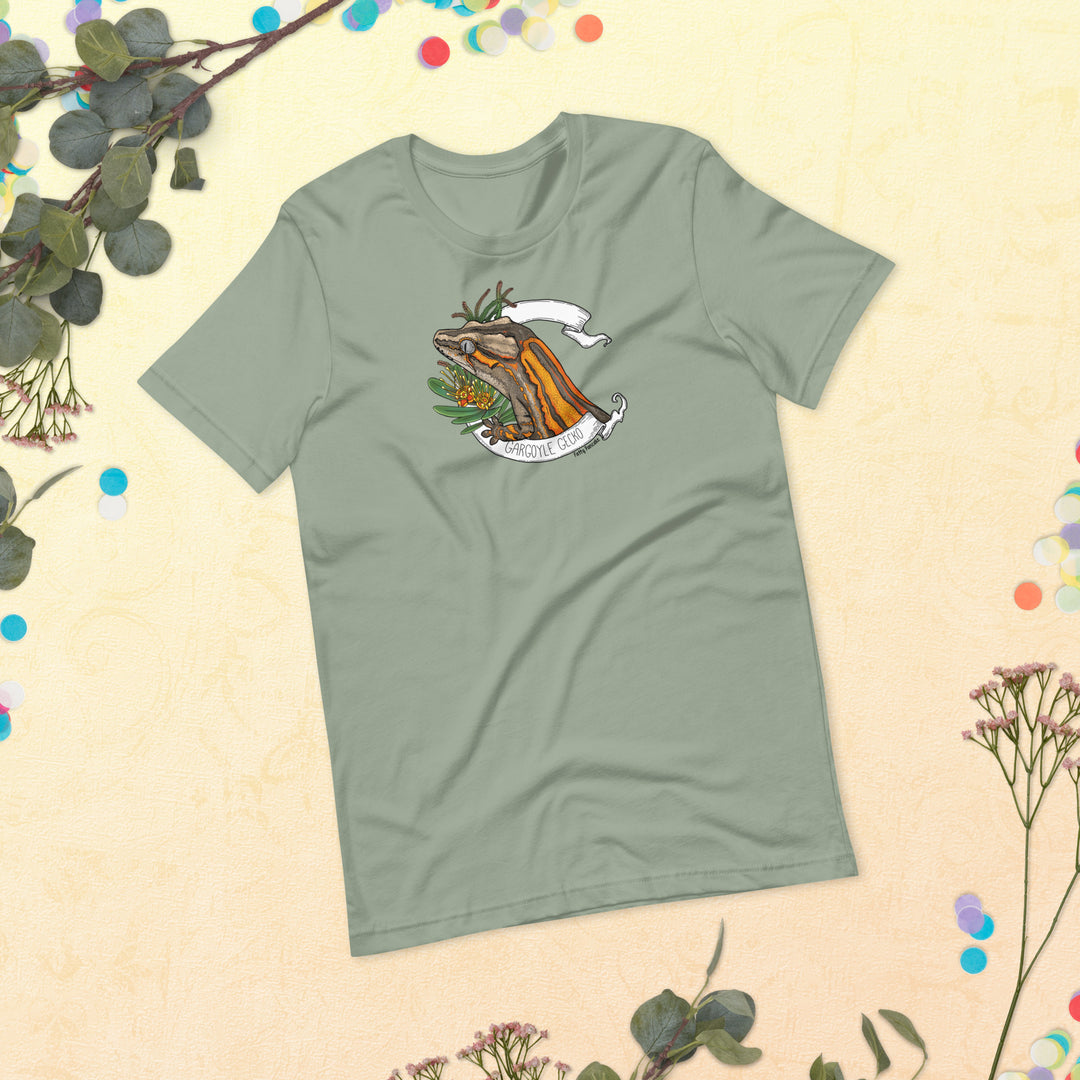 Green t-shirt with a graphic of a gargoyle gecko framed by a white banner and flowers on a beige background with floral elements