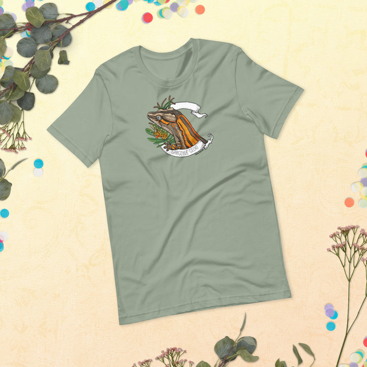 Green t-shirt with a graphic of a gargoyle gecko framed by a white banner and flowers on a beige background with floral elements