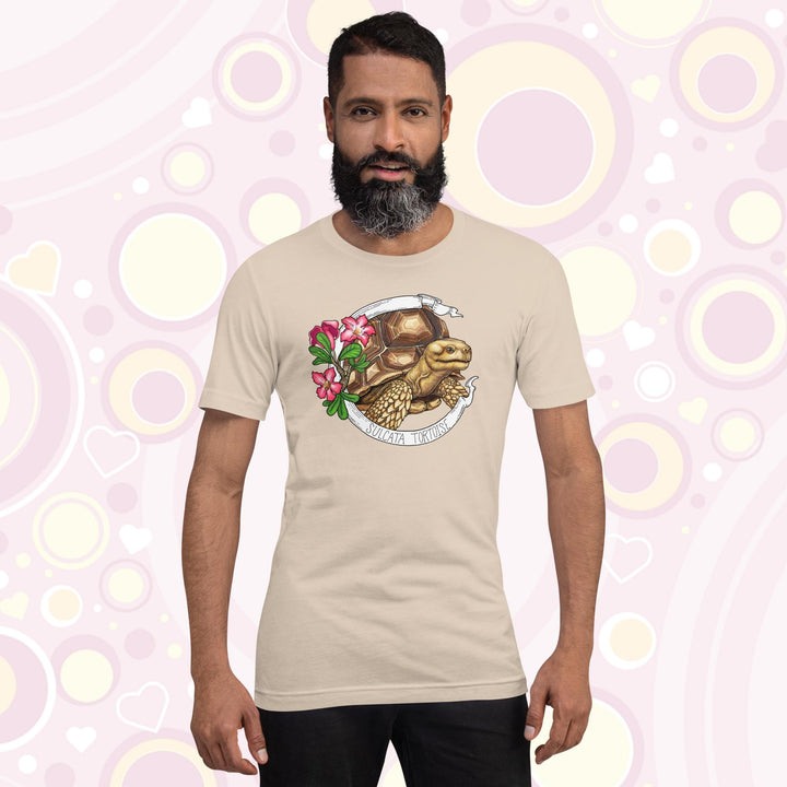 Man wearing soft cream crew neck tee shirt with a sulcata tortoise in the middle of a white banner and pink flowers on the left side of the design. The banner reads Sulcata Tortoise.