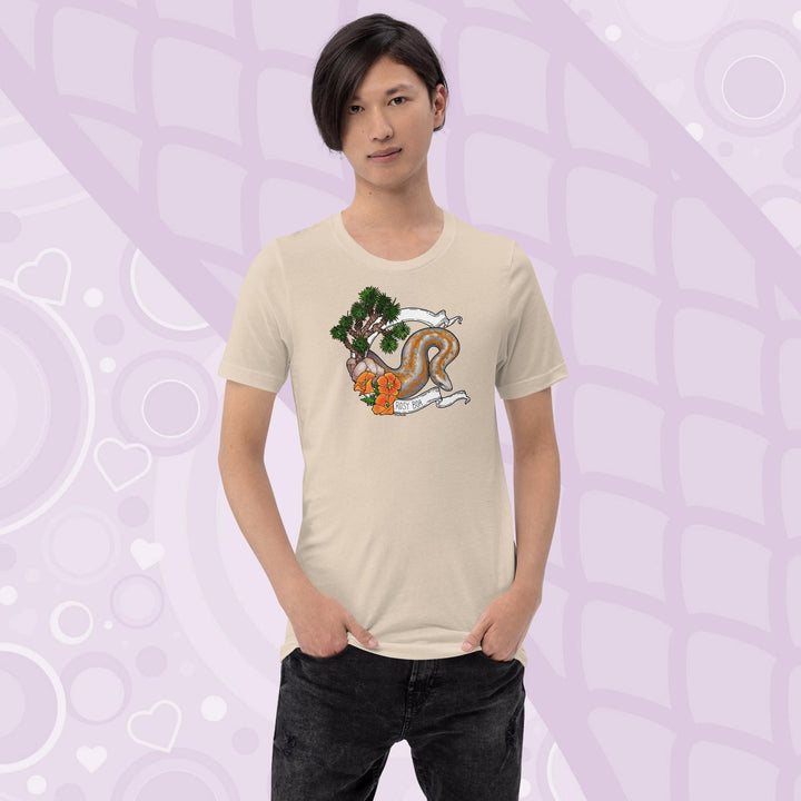 A man wearing a soft cream colored crew cut tee shirt on a light purple background. The art print on the front is a orange and grey speckled rosy boa snake framed by a white banner with California poppy flowers and a Joshua Tree on the left side of the design. The name Rosy Boa is written on the bottom of the banner.