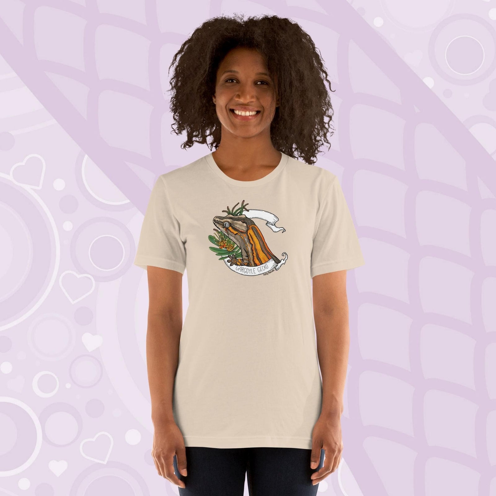 A woman wearing a soft cream colored crew neck shirt with an orange striped gargoyle gecko graphic in the middle of the shirt. 