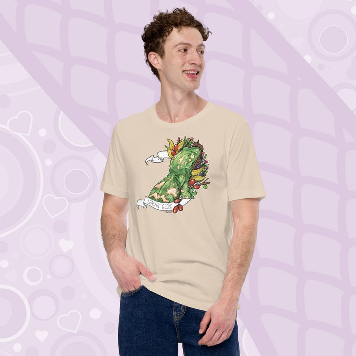 A guy wearing a soft cream colored tee with a green leachie gecko resting on a log and framed by green leaves and red berries. A white banner frames the reptile with the name "Leachie Gecko" on the bottom.