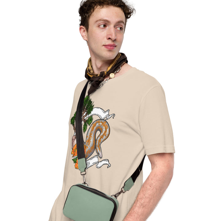 Man wearing a soft cream colored t-shirt with a graphic of the rosy boa printed on it and wearing a green bag, standing against a white background.