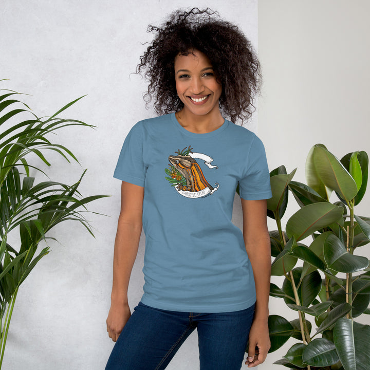Woman wearing a blue t-shirt with a printed graphic of a orange gargoyle gecko, resting between two plants. The woman stands against a white wall with plants behind her.