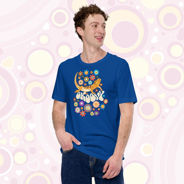 A young man wearing a bright blue crew neck tee showing a groovy bearded dragon design with a 70's retro style with many multi colored flowers surrounding a strutting bearded dragon lizard with the text "groovy" under it's body.