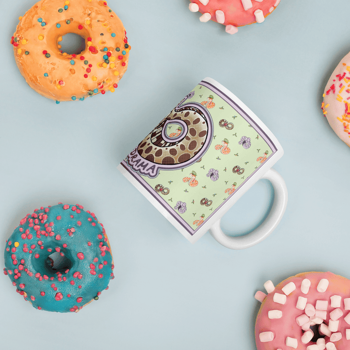 A white ceramic mug with a light green background featuring a repeating pattern of small snakes and a larger central snake graphic with the text "DRAMA" printed in purple. The mug is surrounded by four glazed donuts with colorful sprinkles.