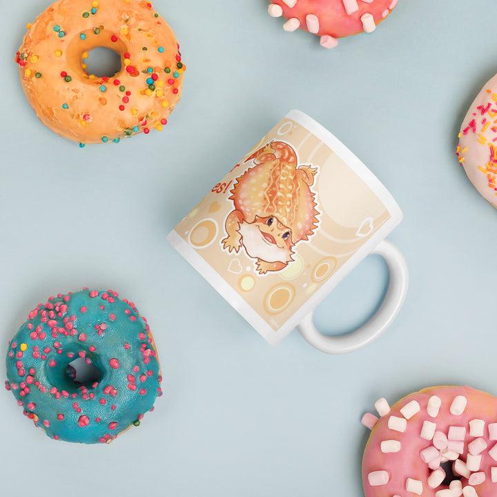 A white ceramic mug with a cute cartoon illustration of an orange colored bearded dragon on a soft cream background, styled in a flat-lay with colorful donuts on a light blue background.