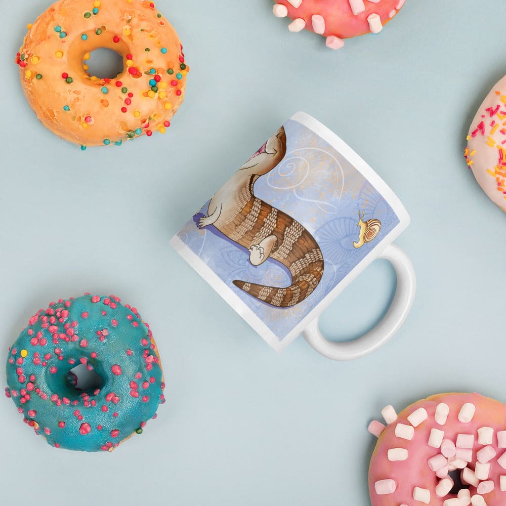 A white ceramic mug with a whimsical illustration of a striped blue tongue skink lizard on a light blue background, styled in a flat-lay with colorful donuts on a light blue surface.