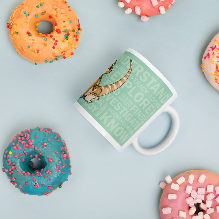 A white ceramic mug featuring a light green background with a graphic of a bearded dragon and the words UNDERSTAND, EXPLORE, INVESTIGATE, and KNOW, surrounded by four colorful donuts with sprinkles and marshmallows.