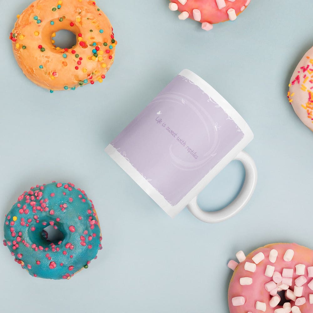 A white ceramic mug with a purple wrap with text that says "Life is sweet with reptiles". It sits on a soft blue background surrounded by donuts glazed in difference colors.