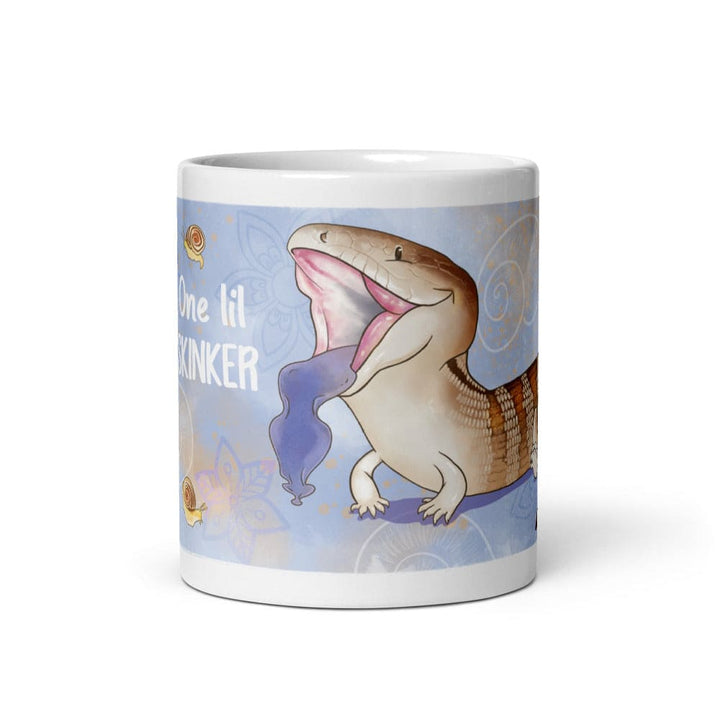 A white ceramic mug featuring a cartoon illustration of a blue-tongued skink with its mouth open, displaying a bright blue tongue. The background is light blue with small snail graphics and the text One lil SKINKER printed in white. The mug sits on a blank white background.
