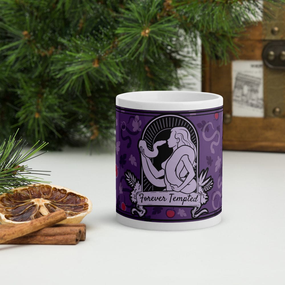 A purple mug with silhouette of woman holding and facing a snake with the text below it reading "Forever Tempted". It sits on a white surface with pine branches in the background.