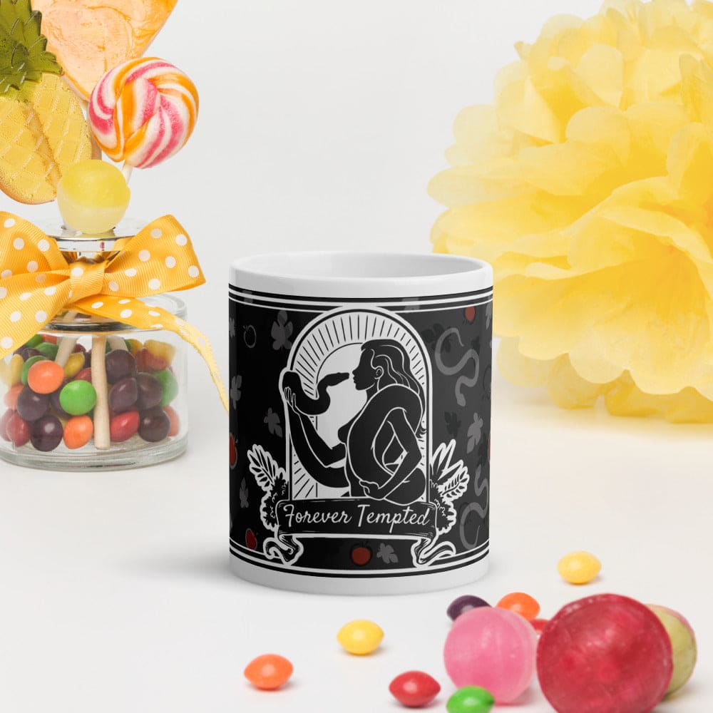 Grey mug with silhouette of woman holding and facing a snake with the text below it reading "Forever Tempted". It sits on a white surface with candy pieces surrounding it.