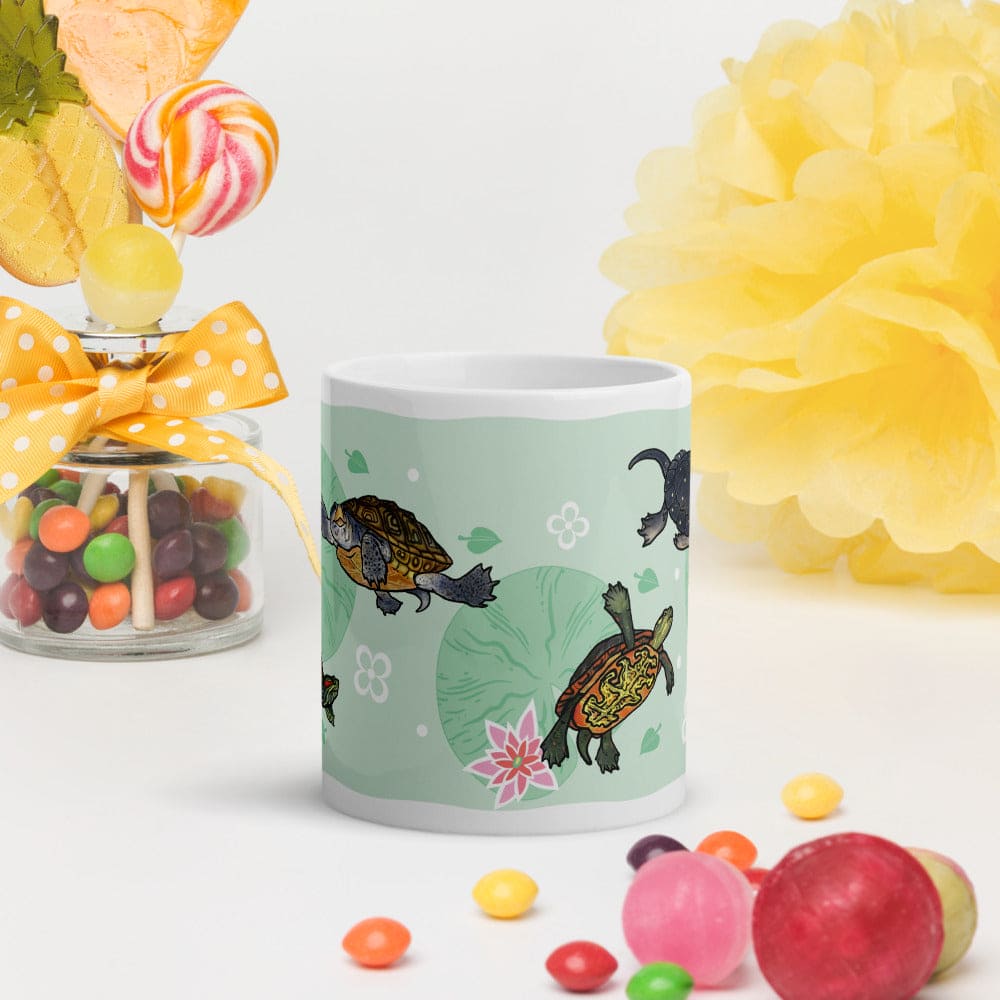 A ceramic mug with a light green design featuring cartoon turtles, lily pads, and a pink flower, styled on a white surface with candy and decorative paper flowers.