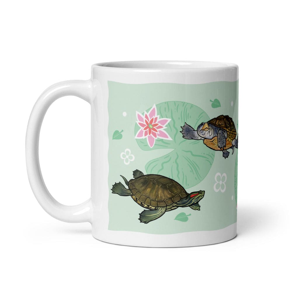 A white ceramic mug featuring a printed design of two turtles on a light green background with a pink lotus flower and lily pad motifs. The handle is on the left side of the mug