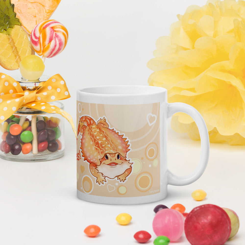 A white ceramic mug with a cute cartoon bearded dragon illustration on a cream and white background, styled with candy and yellow decorative props on a white surface.