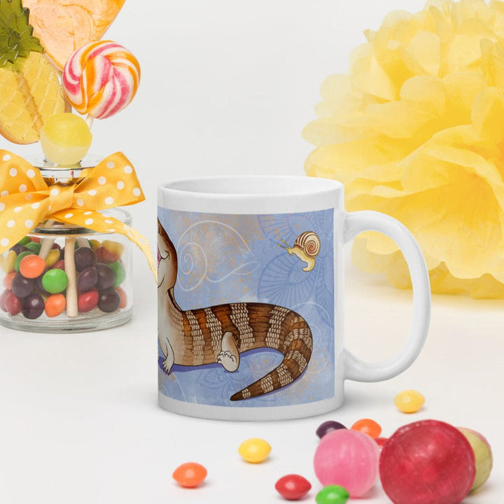 A white ceramic mug with a blue and brown cartoon illustration of a brown blue tongue lizard and a snail, styled on a white surface with candy, a yellow pom-pom, and a decorative jar in the background.