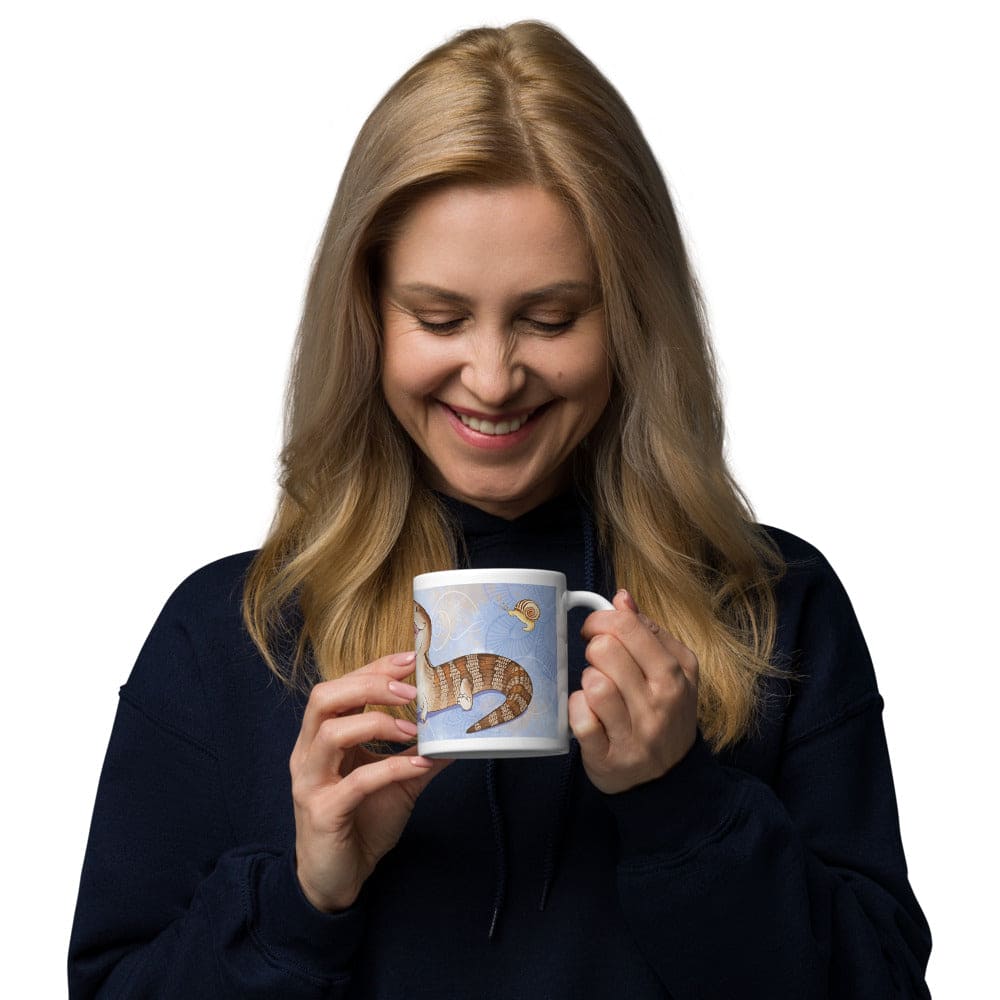 A woman holding a white ceramic mug featuring a detailed illustration of a brown and tan striped lizard with a long tail against a light blue background with faint flower patterns and a small snail graphic near the top right edge of the design.