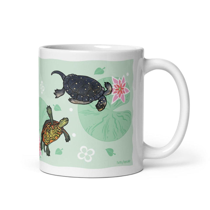 A white ceramic mug featuring a printed design of two turtles on a light green background with a pink lotus flower and lily pad motifs. The handle is on the right side of the mug.