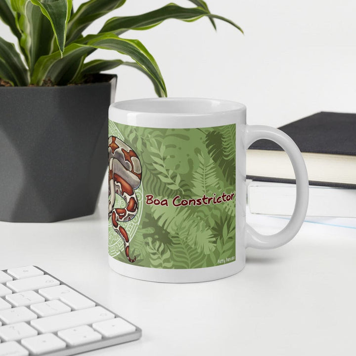 A white ceramic mug with a green tropical leaf pattern and a red-and-white boa constrictor illustration, labeled &