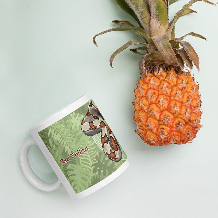 A white ceramic mug featuring a graphic illustration of a red-tailed boa constrictor against a green tropical leaf background with the text Red Tailed printed in red, positioned next to a whole fresh pineapple.