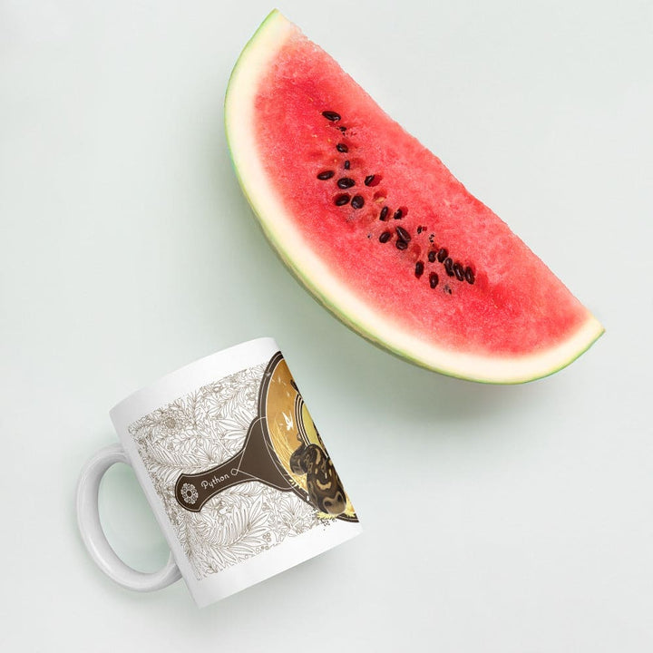 A white ceramic mug featuring a brown and tan graphic of a snake coiled inside a pan, surrounded by a detailed line-art floral pattern and the word Python. A large, fresh slice of red watermelon with black seeds sits nearby.