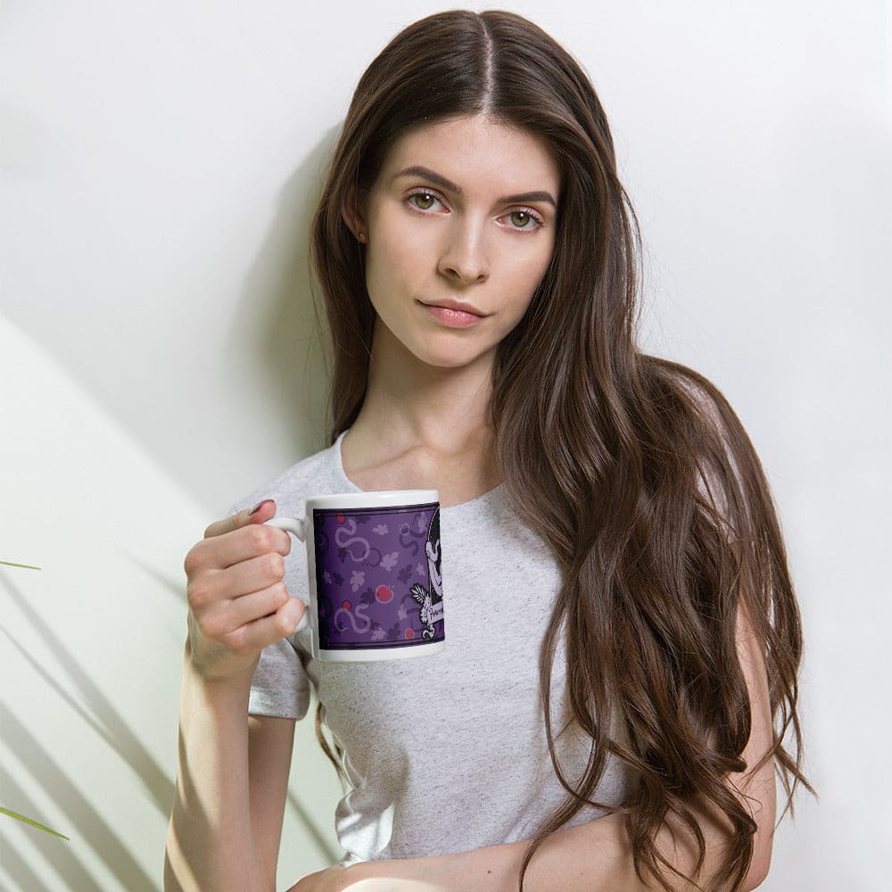 A white ceramic mug featuring a purple background with a repeating pattern of small red apples and gray snake-like shapes. A woman holds the mug while leaning against a white wall.
